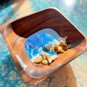 May include: A small wooden bowl with a blue and white resin ocean scene inside. The resin is filled with small white and brown stones.