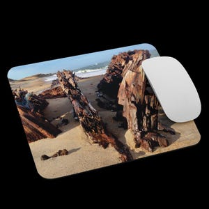 May include: A rectangular mousepad featuring a beach scene with weathered, brown wooden structures, likely remnants of a shipwreck, on a sandy shore. A white computer mouse rests on the right side, against a black background.