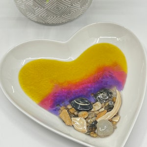 May include: A white heart-shaped dish with a yellow, pink, and purple resin design. The dish is filled with small seashells and stones.