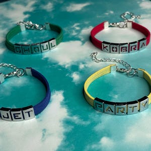 May include: Four colorful bracelets with silver-tone letter beads spelling out "GHOUL", "KOBRA", "JET", and "PARTY". Each bracelet has a different color band: green, red, blue, and yellow. The bracelets are displayed on a light blue background.