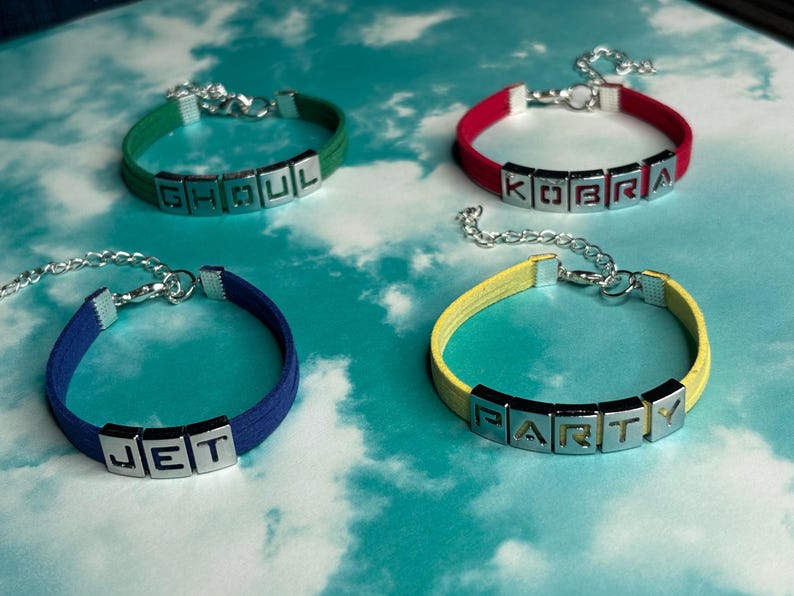 May include: Four colorful bracelets with silver-tone letter beads spelling out "GHOUL", "KOBRA", "JET", and "PARTY". Each bracelet has a different color band: green, red, blue, and yellow. The bracelets are displayed on a light blue background.