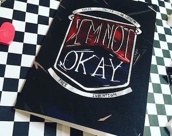May include: A black and white chequered background with a black card featuring a red and white graphic with the text "I'm not okay" in white. The card has Latin text around the graphic.