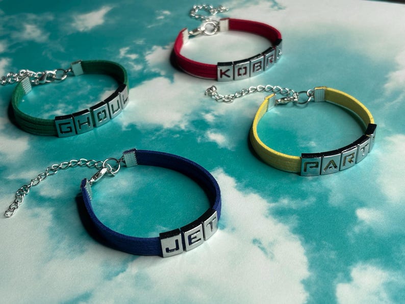 May include: Four colorful bracelets with silver-tone lettered blocks spelling out "GHOU", "KOBR", "PART", and "JET". The bracelets have a simple design with a band in green, red, yellow, and blue. Each bracelet has a silver chain.