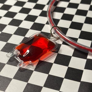 Può includere: Un ciondolo trasparente in plastica a forma di sacca di sangue, riempito di liquido rosso, è appeso a una collana rossa e trasparente. Il ciondolo è rifinito con un anello color argento. Lo sfondo è un motivo a scacchi bianchi e neri.
