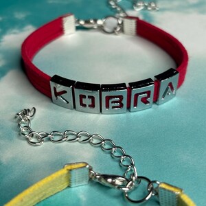May include: A red bracelet with the name "KOBRA" spelled out in silver letter beads. The bracelet has a silver clasp and a matching yellow bracelet is partially visible in the foreground. The background is a light blue and white.