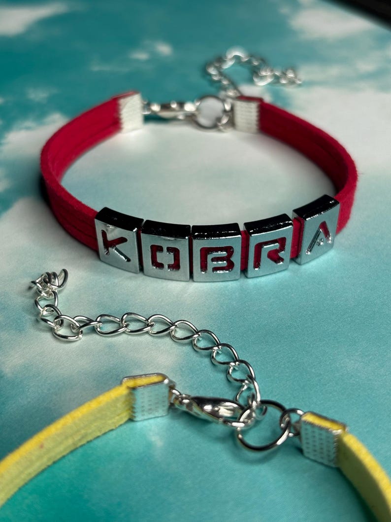 May include: A red bracelet with the name "KOBRA" spelled out in silver letter beads. The bracelet has a silver clasp and a matching yellow bracelet is partially visible in the foreground. The background is a light blue and white.