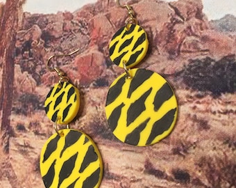 May include: A pair of dangle earrings with two yellow and black circular discs on each earring. The discs have a black and yellow zebra print pattern.