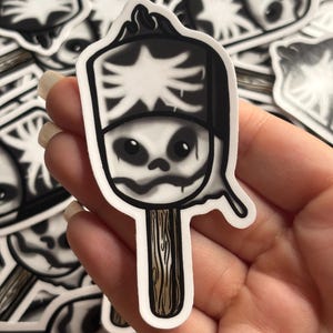 May include: A die-cut sticker shaped like a popsicle with a skull face design. The top portion is black and white with a skeletal rib cage pattern, while the bottom features a brown wooden stick. The overall aesthetic is monochromatic.