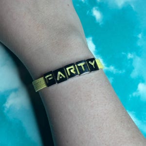 May include: A yellow bracelet with the word "FARTY" spelled out in black and gold letter beads. The bracelet is worn on a wrist against a blue and white background.