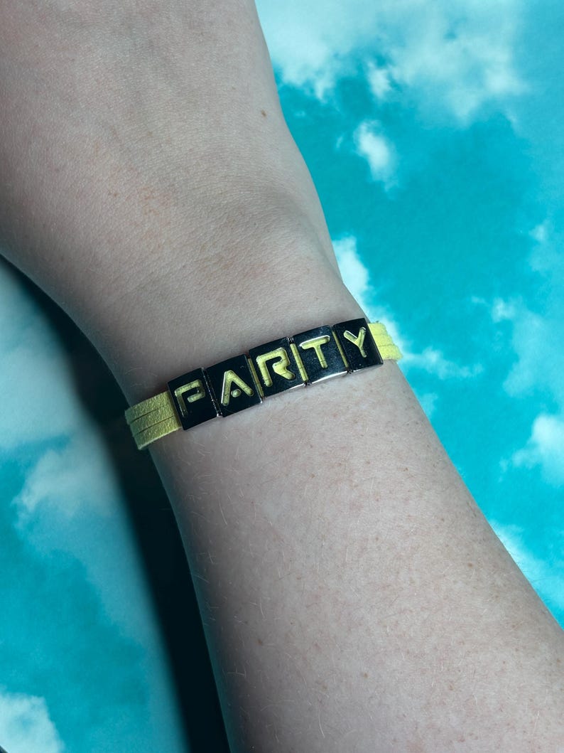 May include: A yellow bracelet with the word "FARTY" spelled out in black and gold letter beads. The bracelet is worn on a wrist against a blue and white background.