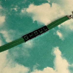 May include: Green suede bracelet with the word "GHOUL" spelled out in black block letters. The bracelet has a silver clasp and is set against a sky-blue background with white clouds.