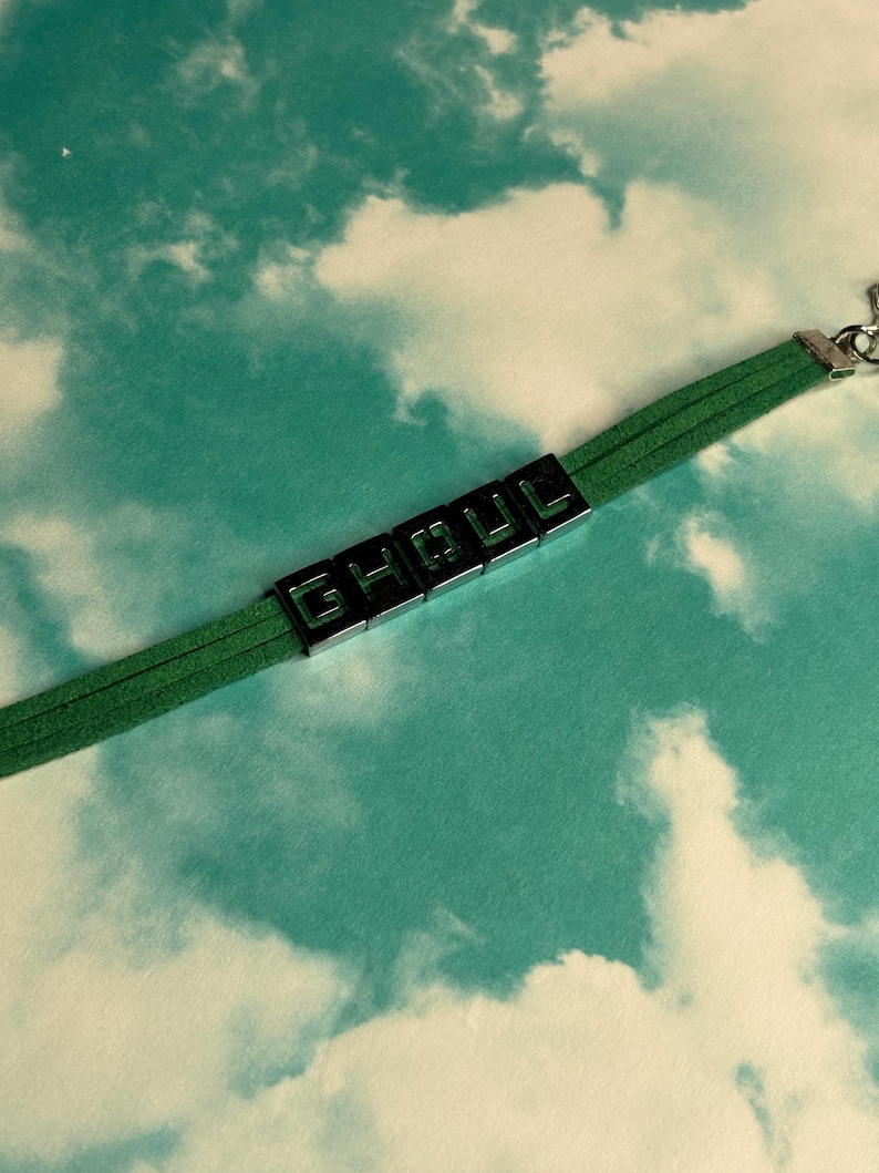 May include: Green suede bracelet with the word "GHOUL" spelled out in black block letters. The bracelet has a silver clasp and is set against a sky-blue background with white clouds.