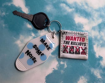 May include: A metal key attached to a split ring with two charms. One charm is a white motel-style tag with blue polka dots and the black text MAKE SOME NOISE. The other is a square charm featuring a newspaper graphic titled Battery City Times.