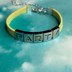 May include: A yellow leather bracelet with silver-tone metal letter beads spelling "PARTY". A red bracelet with the word "KOBRA" is visible in the background. The bracelets are on a blue and white background.