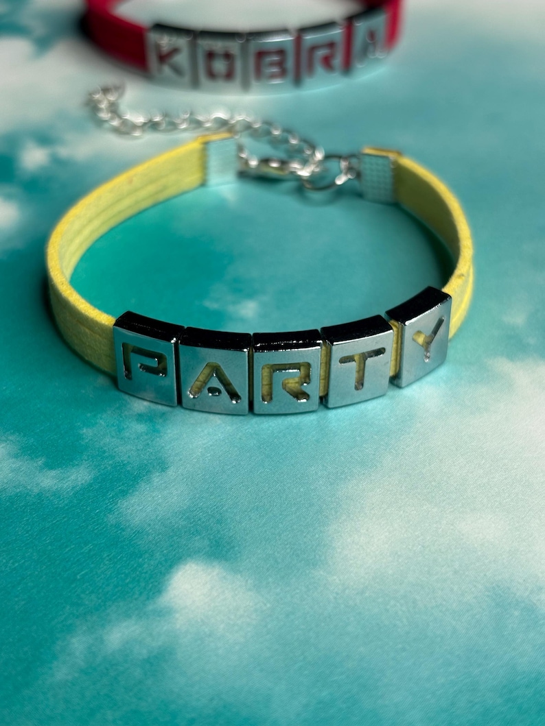 May include: A yellow leather bracelet with silver-tone metal letter beads spelling "PARTY". A red bracelet with the word "KOBRA" is visible in the background. The bracelets are on a blue and white background.