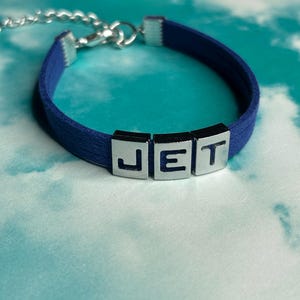 May include: A navy blue bracelet with the letters "JET" in silver block letters. The bracelet has a silver chain clasp. Another bracelet with the letters "GHOUL" is visible in the background.