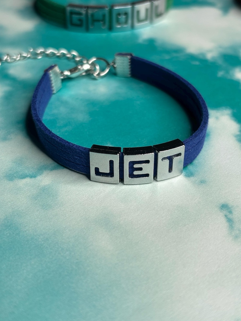 May include: A navy blue bracelet with the letters "JET" in silver block letters. The bracelet has a silver chain clasp. Another bracelet with the letters "GHOUL" is visible in the background.