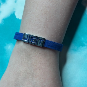 May include: A blue suede bracelet with the word "JET" spelled out in silver metal letters. The bracelet is a simple, minimalist design.