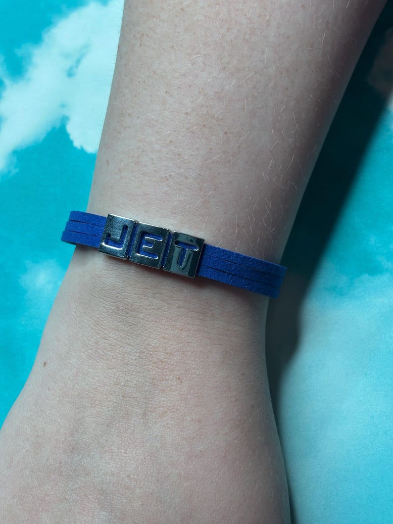 May include: A blue suede bracelet with the word "JET" spelled out in silver metal letters. The bracelet is a simple, minimalist design.