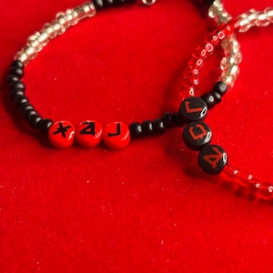 May include: A beaded bracelet featuring a mix of black, clear, and red beads. Three red spherical beads are printed with black symbols, while three black spherical beads are printed with red letters spelling out the word LAC.