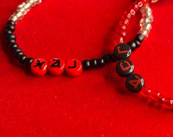 May include: A beaded bracelet featuring a mix of black, clear, and red beads. Three red spherical beads are printed with black symbols, while three black spherical beads are printed with red letters spelling out the word LAC.