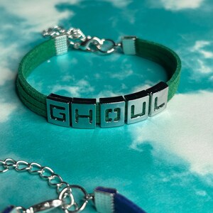 May include: A green suede bracelet with silver-toned metal accents. The bracelet spells out "GHOU L" with individual letter blocks. The bracelet has a silver chain clasp. The background is a light blue and white cloudy pattern.