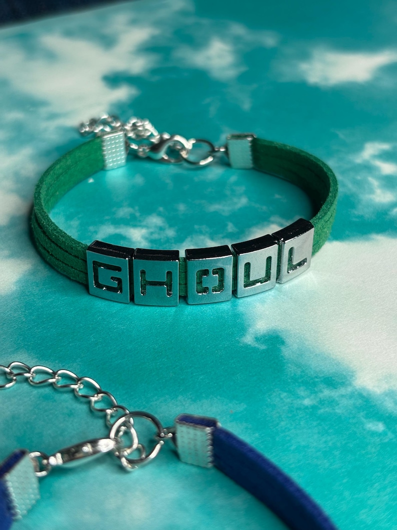 May include: A green suede bracelet with silver-toned metal accents. The bracelet spells out "GHOU L" with individual letter blocks. The bracelet has a silver chain clasp. The background is a light blue and white cloudy pattern.