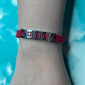 May include: Red suede bracelet with silver metal letter beads spelling "KOBRA". The bracelet is worn on a wrist against a blue and white background. The letters are in a block style.