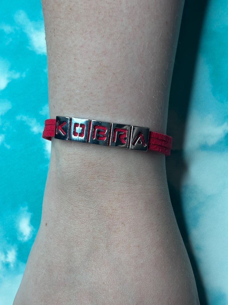 May include: Red suede bracelet with silver metal letter beads spelling "KOBRA". The bracelet is worn on a wrist against a blue and white background. The letters are in a block style.
