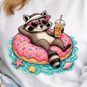 Op de afbeelding: Witte sweatshirt met een cartoon wasbeer die ontspant op een roze donut-drijver in het water. De wasbeer draagt hartvormige zonnebrillen en een vizier, en houdt een drankje vast. De donut heeft hagelslag en de scène bevat schelpen en een zeester.