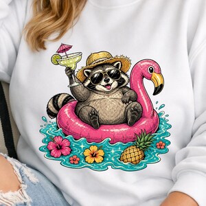 May include: White sweatshirt featuring a cartoon raccoon wearing sunglasses and a straw hat, floating on a pink flamingo pool float. The raccoon holds a cocktail. Tropical flowers and a pineapple are also in the water.