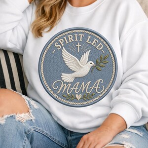 May include: White sweatshirt with a circular embroidered patch. The patch features a dove carrying an olive branch, a cross, and the words "SPIRIT LED MAMA" in a decorative font. The patch is blue with gold and green accents.
