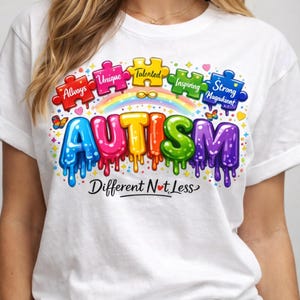 Könnte beinhalten: Weißes T-Shirt mit einem farbenfrohen Grafikdesign. Das Wort "AUTISMUS" wird in leuchtenden, tropfenden Buchstaben dargestellt, darunter der Satz "Different Not Less". Über dem Wort "AUTISMUS" befinden sich Puzzleteile mit Wörtern wie "Always", "Unique" und "Strong".