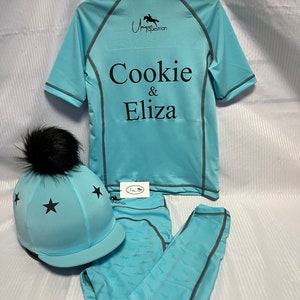 May include: A light blue equestrian set featuring a short-sleeve quarter-zip shirt with the text Cookie & Eliza and a logo, matching leggings with a subtle horse pattern, and a helmet cover with black stars and a black faux fur pom-pom.