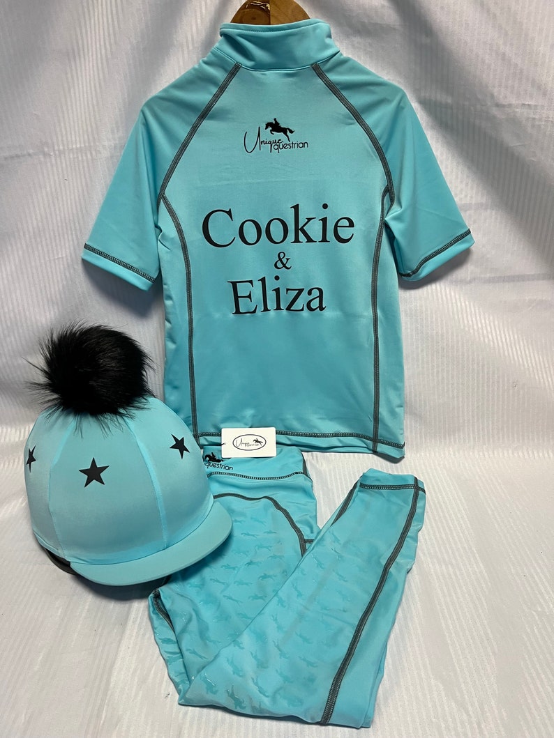 May include: A light blue equestrian set featuring a short-sleeve quarter-zip shirt with the text Cookie & Eliza and a logo, matching leggings with a subtle horse pattern, and a helmet cover with black stars and a black faux fur pom-pom.