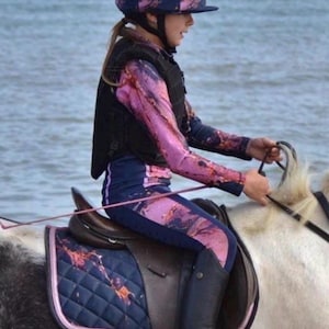 May include: A matching equestrian set featuring a navy blue and pink marble-patterned long-sleeve top, leggings, helmet cover with a pom-pom, and a quilted saddle pad. The set is worn by a rider on a horse, paired with a black protective vest and boots.