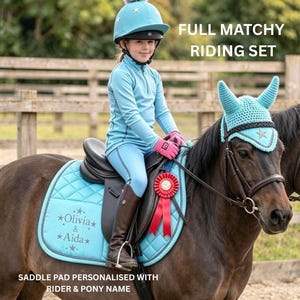 May include: A light blue equestrian riding set featuring a quilted saddle pad personalized with the text Olivia & Aida, a matching crochet ear bonnet with a star emblem, and a light blue riding helmet with a grey pom-pom and star details.