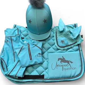 May include: A turquoise equestrian set featuring a helmet with a grey pom-pom and star accents, a quilted saddle pad with the text Unique Equestrian, a matching ear bonnet with a star, a quarter-zip top, and leggings with grey piping details.