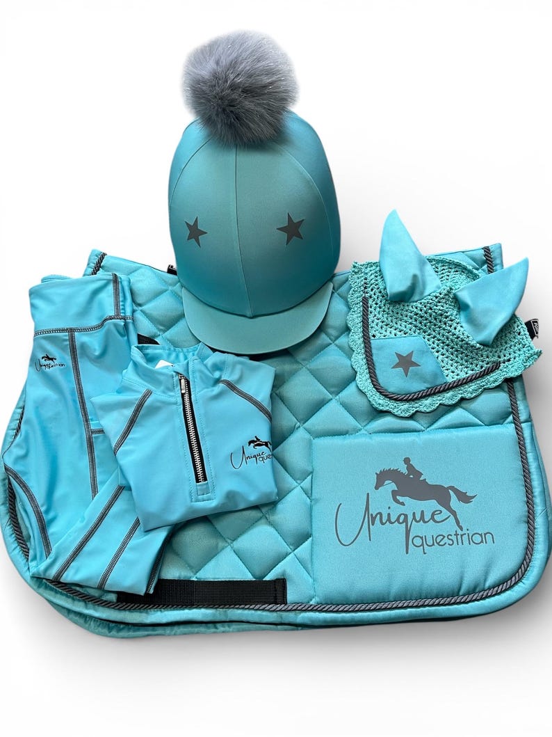May include: A turquoise equestrian set featuring a helmet with a grey pom-pom and star accents, a quilted saddle pad with the text Unique Equestrian, a matching ear bonnet with a star, a quarter-zip top, and leggings with grey piping details.