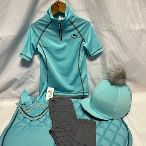 May include: A light blue equestrian set including a short-sleeved quarter-zip shirt with the text Unique Equestrian, a matching saddle pad, a crochet ear bonnet, a grey pair of leggings with a horse pattern, and a helmet cover with a grey pom-pom.