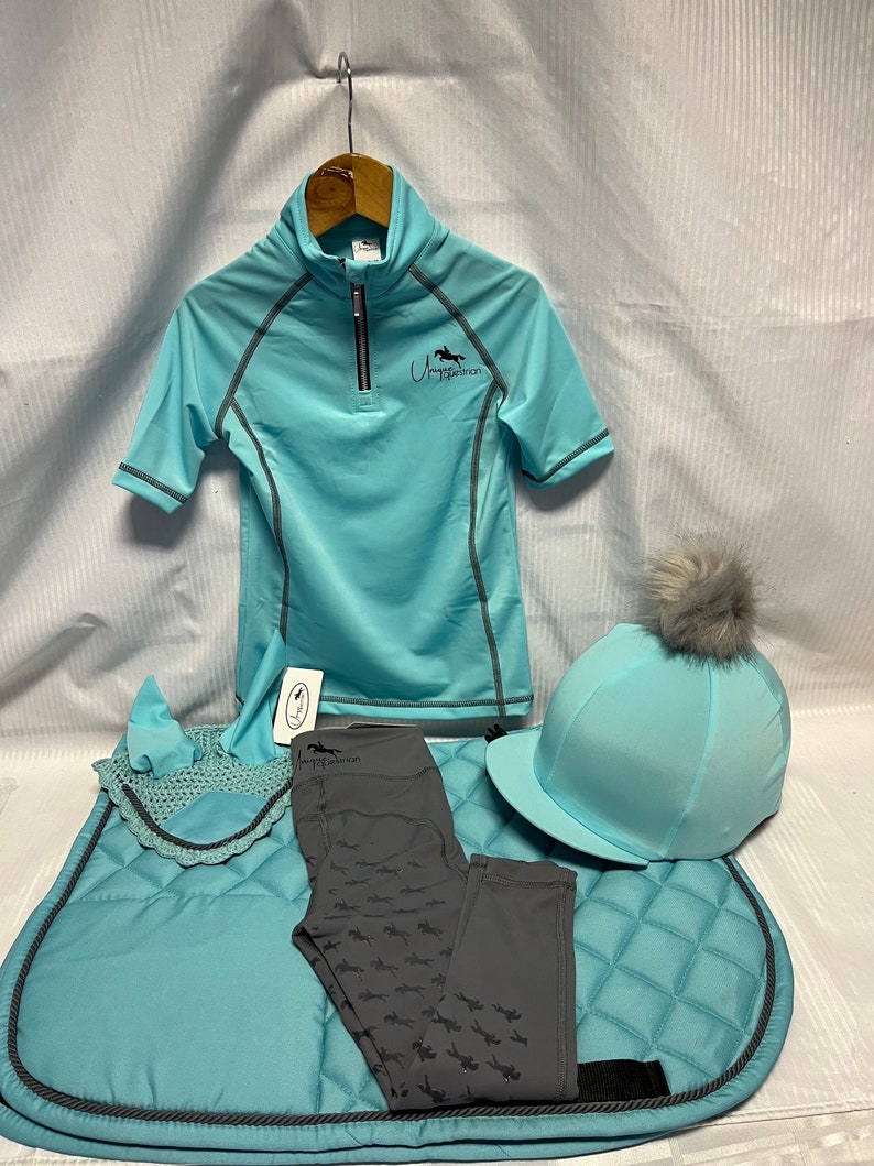 May include: A light blue equestrian set including a short-sleeved quarter-zip shirt with the text Unique Equestrian, a matching saddle pad, a crochet ear bonnet, a grey pair of leggings with a horse pattern, and a helmet cover with a grey pom-pom.