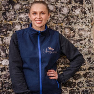May include: A woman wearing a navy blue softshell vest with a blue zipper and a rose gold logo that says "Unique Equestrian" on the front. The vest has a black long-sleeve shirt underneath.