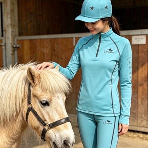 May include: A turquoise equestrian outfit consisting of a long-sleeved quarter-zip top and matching leggings with dark contrast stitching. The set features a small horse logo and text, paired with a matching turquoise baseball cap with star accents.