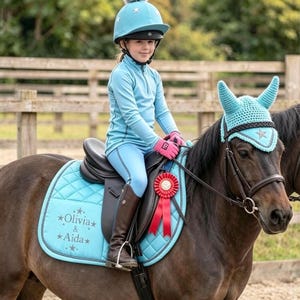 May include: A light blue equestrian set featuring a quilted saddle pad embroidered with the text Olivia & Aida, a matching crochet ear bonnet, and a light blue riding helmet with a gray pom-pom. The set includes a red award ribbon.