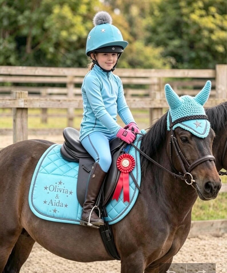 May include: A light blue equestrian set featuring a quilted saddle pad embroidered with the text Olivia & Aida, a matching crochet ear bonnet, and a light blue riding helmet with a gray pom-pom. The set includes a red award ribbon.