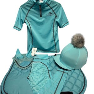 May include: A light blue equestrian set featuring a short-sleeved quarter-zip shirt with a black logo, a quilted saddle pad, a matching ear bonnet, patterned leggings, and a helmet cover with a grey faux fur pom-pom on top of the helmet.