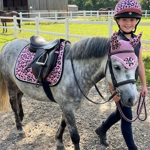 Può includere: Un sottosella e una cuffietta per cavallo abbinata con motivo leopardato rosa e nero. Il sottosella è posizionato sotto una sella in pelle nera su un cavallo grigio, mentre la cuffietta è indossata sulla testa con una corda di guida abbinata.