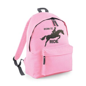 May include: Pink backpack with a black zipper and a front pocket. The backpack features a black graphic of a horse and rider with the words "BORN TO RIDE" printed above and below the image. The straps and top handle are gray.