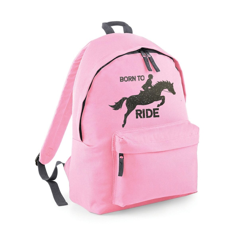 May include: Pink backpack with a black zipper and a front pocket. The backpack features a black graphic of a horse and rider with the words "BORN TO RIDE" printed above and below the image. The straps and top handle are gray.