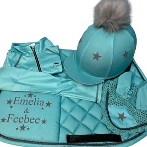 May include: A turquoise equestrian set including a saddle pad, a jacket, a riding hat with a grey pom pom, and a matching neckline. The saddle pad features the text "Emeli & Feebee" with stars.
